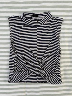 Topshop mörkblå, vit topp, storlek 38 - En topp från Topshop med hög krage och ärmlös design. Toppen har ett randigt mönster i mörkblått och vitt. Den har en omlottdetalj framtill som skapar en draperad effekt som smalnar av precis vid midjan.