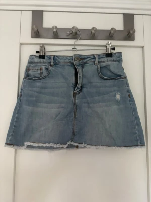 Ljusblå jeanskjol med rå kant - Säljer en ljusblå jeanskjol med rå, fransig kant nertill och slitna detaljer framtill. Kjolen har klassiska fem fickor och bälteshällor i midjan. Perfekt för dig som gillar en avslappnad och trendig stil. Stlk 152 