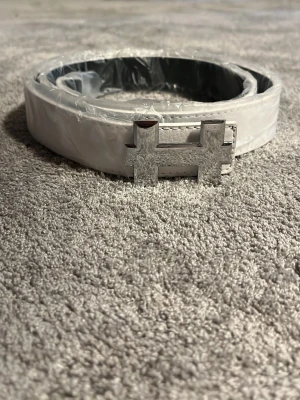 Vitt bälte med silverfärgat H-spänne - Snyggt vitt bälte med ett stort, silverfärgat H-format spänne. Bältet har en clean och modern look, perfekt för att lyfta vilken outfit som helst. Tillverkat i skinn och har en slät yta utan mönster. Passar dig som gillar stilrena accessoarer.