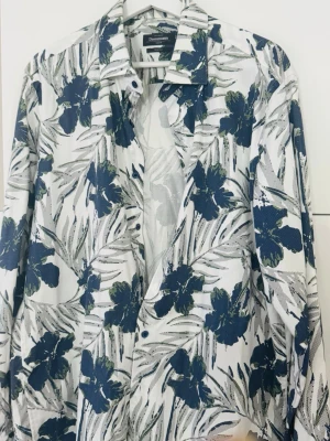 Blommig skjorta från Dressmann - Snygg skjorta från Dressmann med blått och grönt blommönster på vit botten. Skjortan har lång ärm, klassisk krage och knäppning framtill. Slim fit-modell i stretchigt material som sitter snyggt på kroppen.