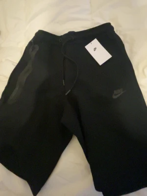 Nike tech shorts - Snygga svarta shorts från Nike med klassisk logga på benet. Perfekta för chill eller träning, tillverkade i mjukt material för skön känsla.