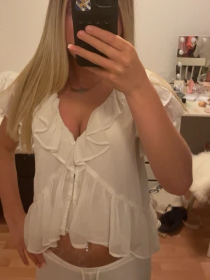 Vit volangblus med knappar - Söt vit blus med volanger och knappar framtill. Blusen har korta ärmar och är gjord i ett lätt, transparent tyg som ger en luftig känsla. Perfekt för en sommardag! Blusen är i storlek XS, den säljs på grund utav att den inte kommer till användning. Köpt för 300 säljs för 150, 50% of!