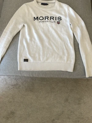 Vit stickad tröja Morris Rowing Club - Vit stickad tröja från Morris Rowing Club med svart broderad logga och klubbmärke på bröstet. Tröjan har rund halsringning, ribbade muddar och nederkant samt en snygg detaljpatch vid fållen. Perfekt för en clean och sportig look. Den är XS men sitter som S