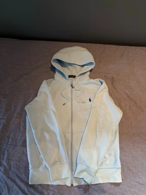 Ralph lauren zip - Snygg ljusblå Ralph lauren zip med dragkedja och huva. Klassisk design med långa ärmar, ribbade muddar och fickor framtill. Liten broderad logga på bröstet. Tillverkad i mjukt bomullsmaterial som är skönt att ha på sig. Perfekt för en avslappnad stil.