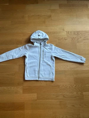 Vit hoodie från Sail Racing - Säljer en vit hoodie från Sail Racing med dragkedja framtill och en extra bröstficka med dragkedja. Hoodien har broderad logga på bröstet och en klassisk huva. Perfekt för dig som gillar stilren och sportig design.