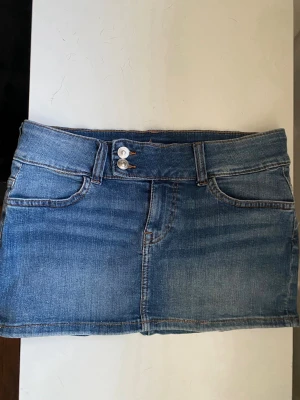  jeanskjol - Snygg blå jeanskjol i klassisk modell med två framfickor och två bakfickor. Kjolen har bälteshällor, dubbla knappar och dragkedja framtill. Perfekt för dig som gillar en enkel och tidlös stil. Storlek 38 men skulle säga att den passar 36 