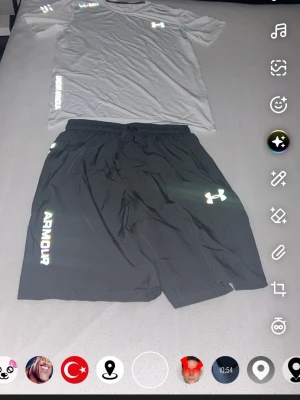 Under Armour träningsset t-shirt & shorts - Säljer ett sportigt träningsset från Under Armour med en vit t-shirt och svarta shorts. T-shirten har tryckt logga och text, och shortsen har elastisk midja samt logga och text på benet. Perfekt för gym eller löpning. Materialet är lätt och andas.