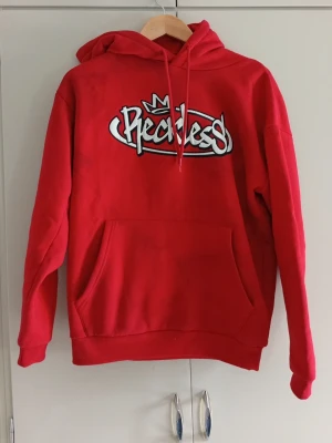 Hoddie  - Snygg röd hoodie från FSBN  med stort vitt och svart tryck på bröstet och en liten krona ovanför loggan. Klassisk känguruficka framtill och justerbar huva med dragsko. Perfekt för dig som gillar streetwear och vill sticka ut lite extra.