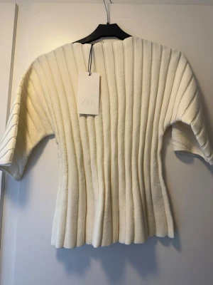 Vit ribbad stickad topp från Zara - Säljer en vit ribbad stickad topp från Zara med korta ärmar och rund hals. Toppen har en figurnära passform och vertikala ribbade detaljer som ger en snygg struktur. Perfekt att styla med jeans eller kjol för en trendig look.