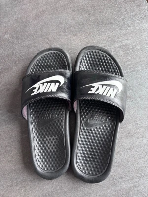 Svarta Nike badtofflor - Säljer ett par svarta Nike badtofflor med vitt logotryck på remmen. Tofflorna har en klassisk slip-on design och mönstrad innersula för extra komfort. Tillverkade i syntetmaterial, perfekta för stranden eller gymmet.
