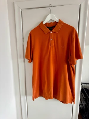 Orange pikétröja från BOSS - Snygg orange pikétröja från BOSS i mjuk bomull. Klassisk krage och två knappar framtill. Perfekt till jeans eller shorts för en avslappnad men stilren look.