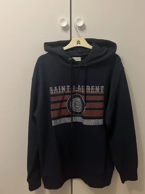 Mörkblå Saint Laurent hoodie - Snygg mörkblå hoodie från Saint Laurent med tryck framtill i vitt och rött samt texten 'SAINT LAURENT'. Hoodien har huva med dragsko och en klassisk känguruficka. Perfekt för dig som vill ha en stilren och trendig look. Storlek S (170-179)