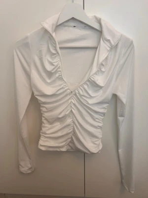 Vit långärmad omlottopp med rynk - Säljer en vit långärmad topp med djup v-ringning och snygga rynkdetaljer framtill. Toppen har en omlottkänsla och är figurnära med stretchigt material. Perfekt att styla med jeans eller kjol för en trendig look.Kan även användas om man ska träna 