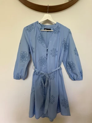 Broderad  sommarklänning  - Ljusblå klänning från Zara med broderade blommönster, knappar framtill och matchande midjebälte. Klänningen har trekvartsärm och lätt v-ringad hals. Använd 1 gång 