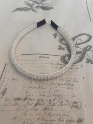 Vitt diadem med pärlor och strass - Snyggt vitt diadem dekorerat med små pärlor och glittrande strassdetaljer. Diademet har en klassisk form och är vadderat på insidan för extra komfort. Perfekt accessoar för att lyfta din look med lite extra bling.