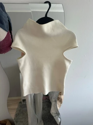 Vit ärmlös polotopp - Säljer en stilren vit polotopp i tjockare material. Toppen är ärmlös och har en hög krage som ger en modern look. Perfekt att styla med jeans eller kjol för en trendig outfit.
