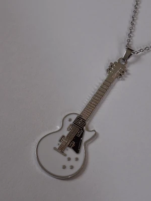 Halsband med gitarrhänge i silverfärg - Unikt halsband med ett hänge en elgitarr i silverfärgad metall och vit emalj. Kedjan är tunn och smidig, och gitarrhänget har detaljer som strängar och knappar. Materialet är rostfritt stål. 