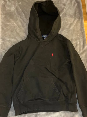 Mörkgrön hoodie från Polo Ralph Lauren - Mörkgrön hoodie från Polo Ralph Lauren med klassisk huva och känguruficka. På bröstet finns den ikoniska röda Polo-loggan broderad. Hoodien har ribbade muddar vid ärmslut och nederkant. Perfekt för en avslappnad och stilren look. Vid frågor hör av er och priset kan alltid diskuteras 
