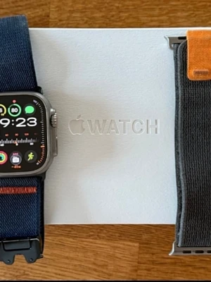Apple Watch Ultra med blått band - Apple Watch Ultra med robust fyrkantig titanboett och en stor, tydlig skärm. Klockan har ett blått textilband med orange detaljer och svart spänne. Perfekt för dig som vill ha en smartklocka med sportig design och avancerade funktioner.
