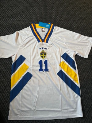 Svensk landslagströja Adidas nr 11 - Vit svensk landslagströja från Adidas med nummer 11, blå och gula detaljer samt SVFF-emblem på bröstet. Tröjan har V-ringad krage med blå, gul och röd rand. Perfekt för dig som älskar fotboll och vill stötta Sverige.