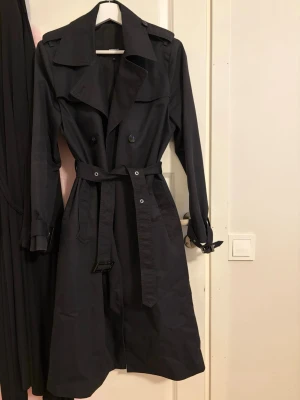 Svart klassisk trenchcoat med bälte - Stilren svart trenchcoat med dubbelknäppning, bred krage och avtagbart midjebälte. Jackan har axelklaffar, justerbara ärmslut och slät finish. Perfekt för dig som gillar tidlös och elegant stil. Materialet känns som bomullsmix. Strl 40 men passar även 36/38