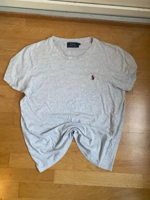 Grå t-shirt från Polo Ralph Lauren - Klassisk ljusgrå t-shirt från Polo Ralph Lauren med korta ärmar och rund hals. T-shirten har den ikoniska vinröda broderade loggan på bröstet. Tillverkad i mjuk bomull för en skön och avslappnad passform.