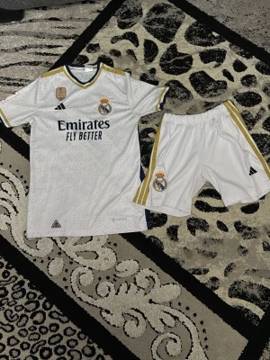 Real Madrid matchställ Bellingham #5 - Säljer ett vitt Real Madrid matchställ från Adidas med Bellingham #5 på ryggen och shorts. Tröjan har klubbmärke, sponsortryck och gulddetaljer på axlarna. Shortsen är vita med guldränder och klubbmärke. Perfekt för fotbollsträning eller match!