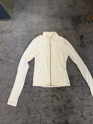 Vit sportjacka från Gina Young - Vit sportig jacka från Gina Tricot med hel dragkedja framtill och hög krage. Tillverkad i ett stretchigt syntetmaterial som ger en tight och smickrande passform. Perfekt för träning eller som ett extra lager under kyligare dagar.