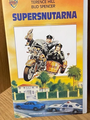 Supersnutarna VHS-film - Supersnutarna är en klassisk VHS-film med färgstarkt omslag i orange och gult. Framsidan har en tecknad bild av två poliser på motorcykel och bilar. Filmen är svensktextad och har Terence Hill och Bud Spencer i huvudrollerna. Perfekt för dig som gillar retro och samlar på gamla filmer.