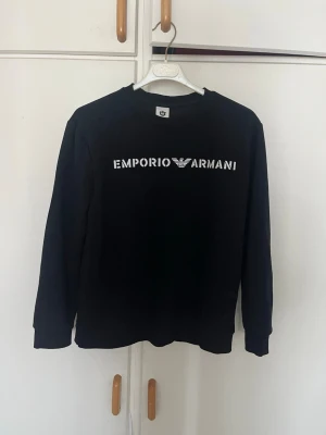 Svart sweatshirt från Emporio Armani pris kan diskuteras - Stilren svart sweatshirt från Emporio Armani med vit logotyp och text på bröstet. Tröjan har rund halsringning och långa ärmar. Perfekt för en avslappnad och trendig look.