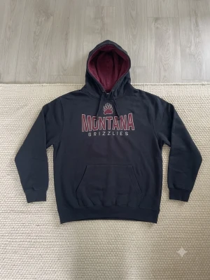 Svart Montana Grizzlies hoodie XL - Snygg svart hoodie från Stadium Athletics med trycket 'Montana Grizzlies' och björntass på bröstet. Hoodien har vinrött foder i huvan och en stor magficka. Perfekt för dig som gillar college-stil och vill ha en bekväm och cool look.