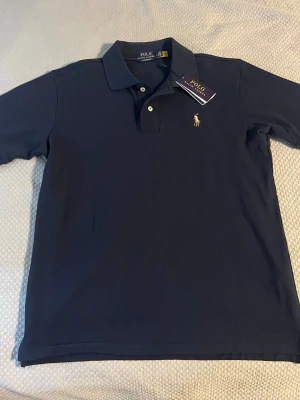 Mörkblå pikétröja från Polo Ralph Lauren - Klassisk mörkblå pikétröja från Polo Ralph Lauren med krage, knappar och den ikoniska broderade loggan på bröstet. Perfekt för en stilren och avslappnad look. Tillverkad i mjuk bomull för extra komfort.