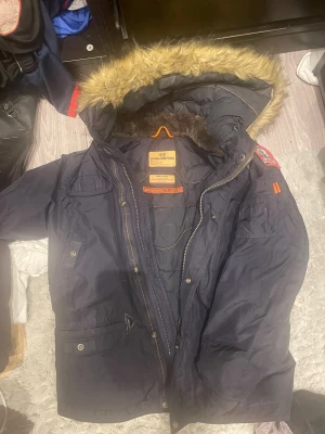 Mörkblå parkas från Parajumpers - Snygg mörkblå parkas från Parajumpers med avtagbar huva och fuskpäls. Jackan har flera fickor, dragkedja och tryckknappar samt detaljer i orange. Perfekt för kalla dagar och riktigt varm med mjukt innerfoder.