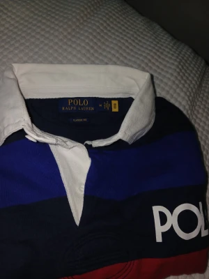 Randig långärmad skjorta Polo Ralph Lauren - Färgglad långärmad skjorta från Polo Ralph Lauren med breda ränder i blått, rött, grönt, gult och orange. Klassisk vit krage och stor POLO-logga på bröstet. Perfekt för dig som gillar statement-plagg och sportig stil.