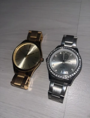 Guld & Silver Klocka - Säljer två snygga klockor från Regal. Den ena är helguld med minimalistisk urtavla och länkarmband, den andra är silvrig med glittrande stenar runt boetten och länkarmband. Båda har klassisk rund form och passar dig som gillar stilren design.