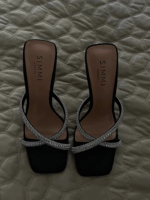 Svarta pumps med strass från ASOS - Säljer ett par svarta pumps från ASOS med fyrkantig tå och höga klackar. Skorna har två korsade remmar täckta av glittrande strass som ger en lyxig känsla. Materialet är en mix av textil och syntet. Perfekta för dig som vill sticka ut.