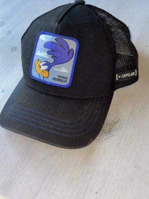 Svart keps Capslab Road Runner - Snygg svart keps från Capslab med Road Runner-motiv framtill. Kepsen har mesh på baksidan för extra ventilation och justerbar passform. Framtill finns en broderad patch med tecknad Road Runner och blå detaljer på skärmen. Säljer den eftersom jag har fått en annan keps