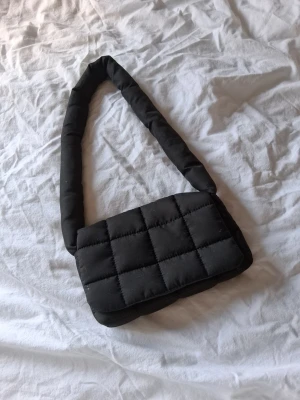 Svart quiltad axelväska - Trendig svart handväska med quiltat mönster och vadderad axelrem. Väskan har magnetlås och är tillverkad i ett mjukt tygmaterial. Perfekt för dig som gillar en stilren och modern look.