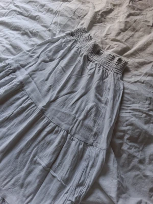 Vit långkjol med smock - Långkjol i vit med bred smock i midjan och volangdetaljer. Kjolen har en luftig och flowy passform, perfekt för varma dagar. Tillverkad i ett mjukt och lätt tyg som ger en avslappnad känsla.