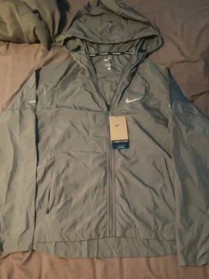 Grå vindjacka med huva från Nike - Snygg grå vindjacka från Nike med huva och dragkedja framtill. Jackan har två sidofickor med dragkedja och en vit Nike-logga på bröstet. Tillverkad i lätt och vindtätt material, perfekt för blåsiga dagar.