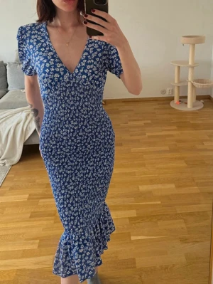Blå blommig långklänning med volang - Snygg blå långklänning med vitt blommönster och volang nertill. Djup v-ringning och kortärmad modell som sitter figurnära. Perfekt för dig som gillar romantisk och feminin stil.