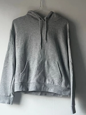 Grå basic hoodie med ficka - En enkel och stilren grå hoodie med huva och dragsnören. Hoodien har en stor magficka framtill och ribbade muddar vid ärmslut och nederkant. 