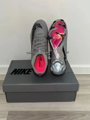 Nike grå fotbollsskor Air Zoom - Snygga grå elite  Nike Air Zoom fotbollsskor med svart mönster och detaljer. Skorna har en låg profil, snörning och en cool holografisk sula med rosa inslag. Perfekta för dig som vill sticka ut på planen och få bra grepp och känsla i spelet.