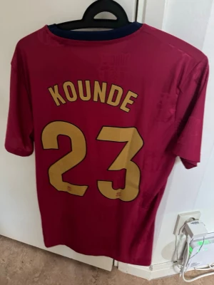 Barcelona matchtröja Kounde 23 - Säljer en vinröd FC Barcelona matchtröja från Nike med Kounde och nummer 23 på ryggen. Tröjan har klubbmärke och sponsorlogga på bröstet samt mörkblå detaljer vid kragen. Perfekt för dig som älskar fotboll och vill stötta Barca.