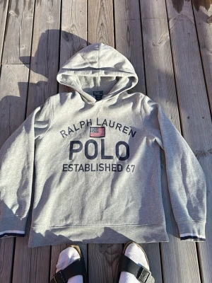 Grå hoodie från Ralph Lauren Polo - Snygg grå hoodie från Ralph Lauren Polo med stor logga och amerikansk flagga på bröstet. Klassisk huva och långa ärmar. Mjuk bomullskänsla och avslappnad passform, perfekt för en chill och sportig stil. OBS barnstorlek 