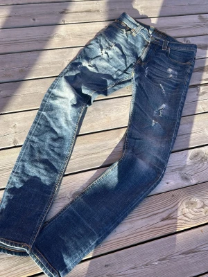 Blå slitna jeans med hål - Säljer ett par blå jeans med slitningar och hål för en riktigt avslappnad och trendig look. Jeansen har klassisk femficksmodell, normal midja och raka ben. Materialet är denim med snygg tvätt och slitdetaljer framtill.