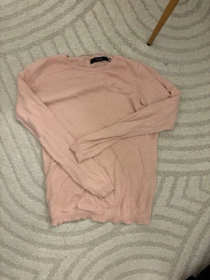 Ljusrosa stickad tröja Vero Moda - Söt ljusrosa stickad tröja från Vero Moda med diskret hålmönster. Tröjan har rund halsringning, långa ärmar och ribbade muddar vid ärmslut och nederkant. Perfekt för lager på lager och enkel att matcha med jeans eller kjol.