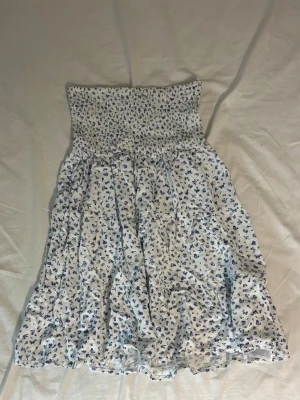 Vit blommig sommarklänning - Söt vit kortklänning med blått blommönster och smock upptill. Axelbandslös modell med luftig kjol, perfekt för varma dagar. Tillverkad i mjukt bomullstyg som känns skönt mot huden.