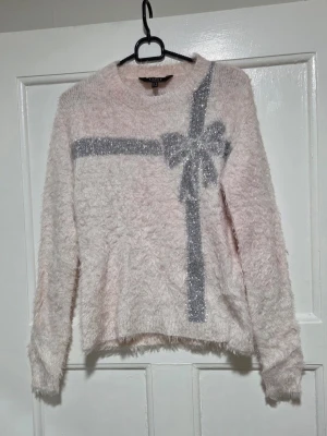 Ljusrosa fluffig tröja med rosett - Supersöt ljusrosa stickad tröja från Lipsy med fluffig textur. Tröjan har ett glittrigt silvermönster som ser ut som ett paketband med en stor rosett framtill. Perfekt för dig som gillar gulliga och unika plagg. Rund hals och långa ärmar.
