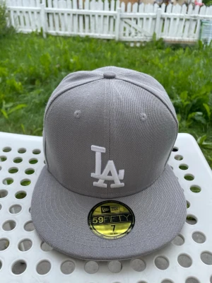 Grå LA 59FIFTY keps från New Era - Säljer en stilren grå 59FIFTY keps från New Era med LA-logga broderad i vitt framtill. Klassisk flat brim och strukturerad passform. Kepsen är tillverkad i ett slitstarkt tyg och har officiella etiketter kvar. Perfekt för dig som gillar streetwear och sportig stil.